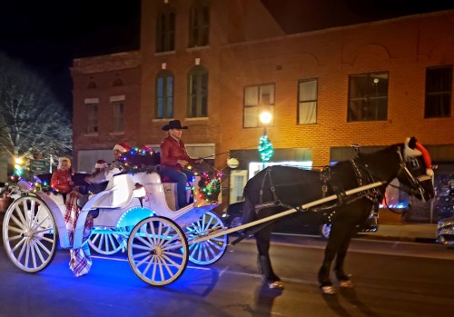 A decorated horse-drawn carriage with riders in festive attire, illuminated by colorful lights, travels down a street at night.