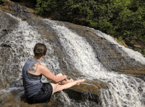 A person sits on a rocky ledge by a waterfall, enjoying the natural scenery and splashing water.