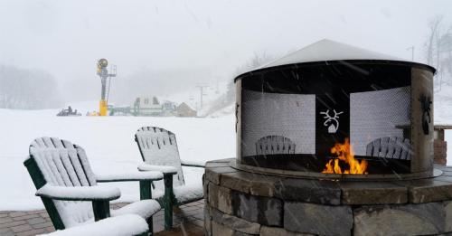 A snowy scene featuring a fire pit with chairs, a ski lift in the background, and a cozy winter atmosphere.