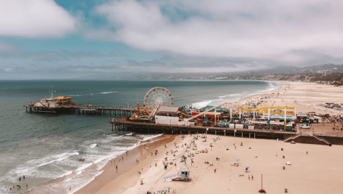 Aerial view of a beach with a pier, amusement park, and people enjoying the sandy shore under a cloudy sky.