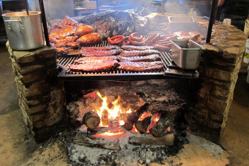 A smoky barbecue grill with various meats, including ribs and sausages, cooking over an open flame.