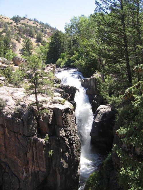 A waterfall cascades between rocky cliffs, surrounded by lush green trees and a mountainous landscape.