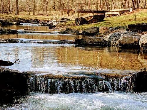 A serene stream flows over rocks, surrounded by trees, with a wooden bridge in the background.