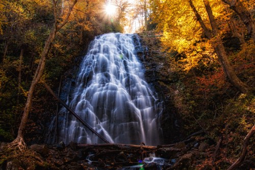 A cascading waterfall surrounded by vibrant autumn foliage and sunlight filtering through the trees.