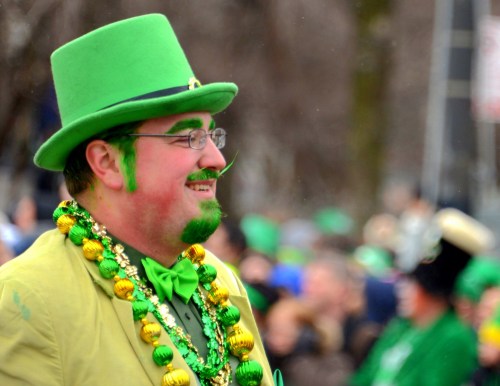 A man in a green top hat and suit, adorned with beads, smiles during a festive parade celebration.