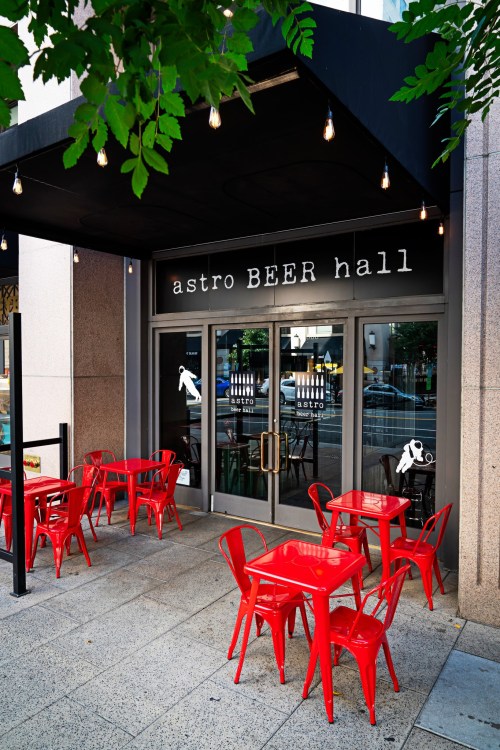 Exterior of Astro Beer Hall featuring red tables and chairs under string lights, with large glass doors.
