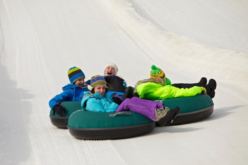 Four children in colorful winter gear joyfully sledding down a snowy hill on inflatable tubes.