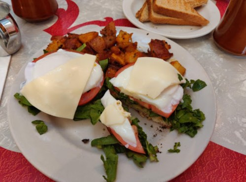 A plate with two eggs Benedict on toast, topped with cheese, spinach, and tomatoes, served with roasted potatoes.
