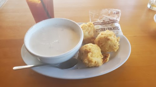 A bowl of creamy soup with three golden biscuits and a packet of oyster crackers on a white plate.