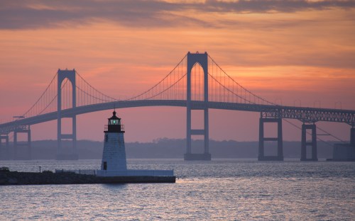 A lighthouse stands in the foreground with a bridge and sunset in the background, casting a serene atmosphere over the water.