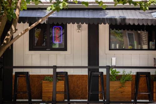 A cozy café exterior with an "OPEN" neon sign, black stools, and planters under a shaded awning.
