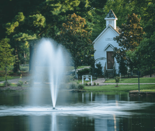 A serene pond with a fountain, surrounded by trees, featuring a small white chapel in the background.