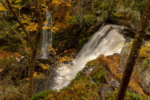 A serene waterfall cascades over rocks, surrounded by autumn foliage and trees in a tranquil forest setting.