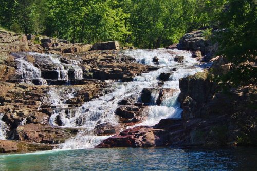 A serene waterfall cascading over rocky terrain, surrounded by lush green trees and a clear blue pool below.