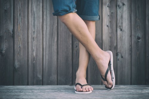 A person wearing flip-flops stands with crossed legs on a wooden surface against a rustic wooden background.