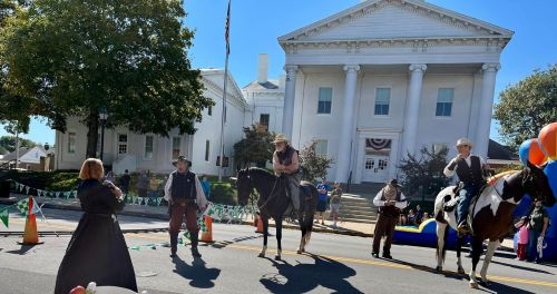 A parade scene with people on horseback in front of a historic building, decorated with festive banners and balloons.