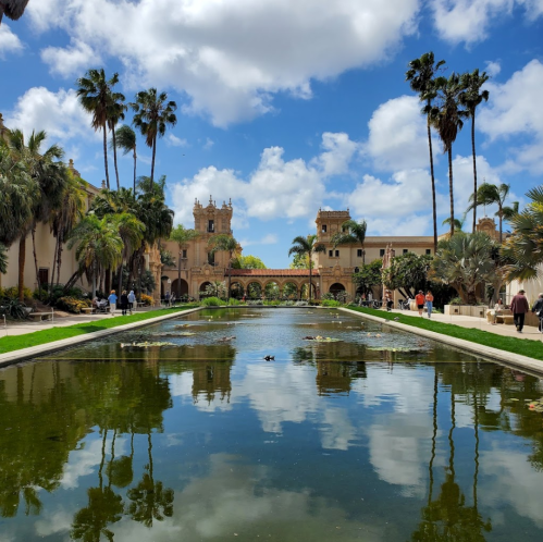 A serene park scene featuring a pond reflecting palm trees and historic buildings under a blue sky with fluffy clouds.