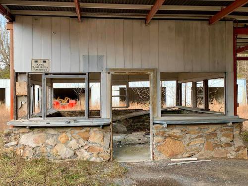 Abandoned building interior with stone counter, empty windows, and overgrown grass outside. Sign reads "Keep Seat Belts Fastened."