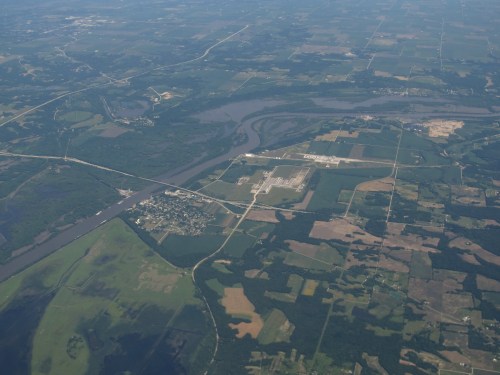 Aerial view of a river winding through green fields and farmland, with a small town and industrial area visible.
