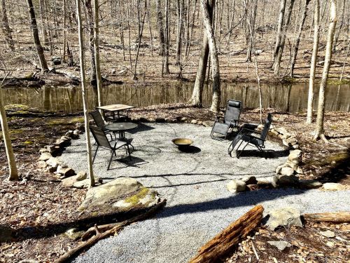 A serene outdoor seating area with chairs and a table, surrounded by stones, near a calm stream in a wooded setting.