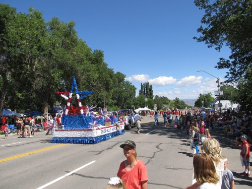 A festive parade scene with a decorated float and crowds of people lining the street under a clear blue sky.