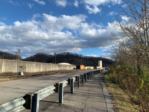 A paved road lined with a guardrail, leading to a distant hill under a partly cloudy sky.