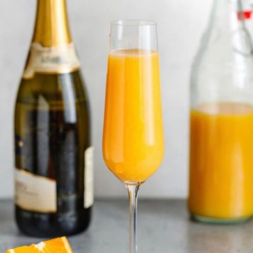 A glass of orange mimosa with champagne bottles and a jug of orange juice in the background.