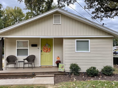 A cozy beige house with a green door, two chairs on the porch, and autumn decorations in the front yard.