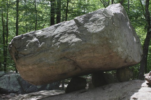 A large, balanced rock resting on smaller stones in a forested area with green trees in the background.