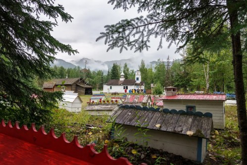A serene landscape featuring small, colorful wooden structures and a church, surrounded by lush greenery and mountains.