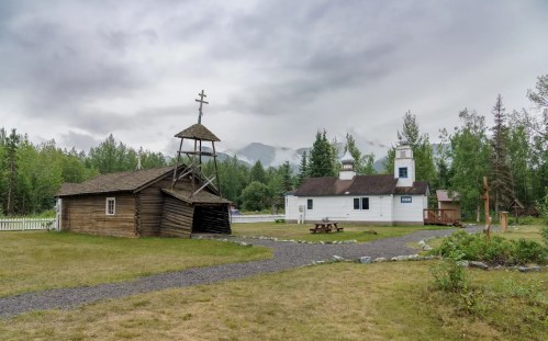 A rustic wooden church and a white building surrounded by trees under a cloudy sky, with a gravel path and picnic area.