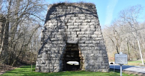 Stone structure with an archway, surrounded by trees, with a historical marker nearby. Clear blue sky above.