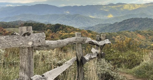 A rustic wooden fence overlooks a colorful mountain landscape with rolling hills and a cloudy sky.