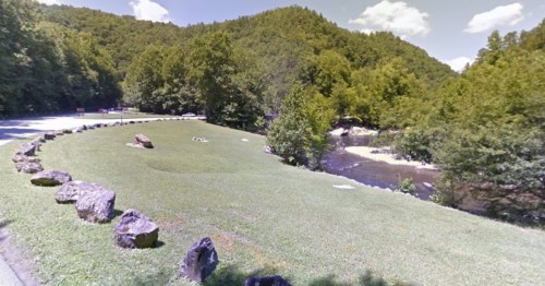 A grassy area with rocks, surrounded by trees and a river, set against a backdrop of rolling hills and blue sky.