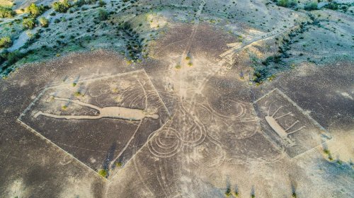 Aerial view of large geoglyphs etched into a dry landscape, featuring abstract shapes and patterns.