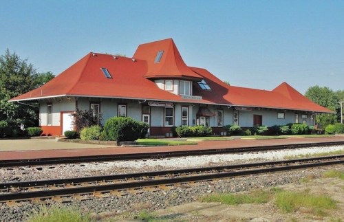 Historic train station with a distinctive red roof, surrounded by greenery and railway tracks.