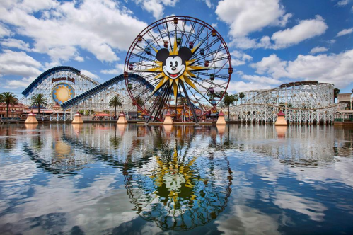 A colorful amusement park scene featuring a large Ferris wheel with a Mickey Mouse design, reflecting in the water.