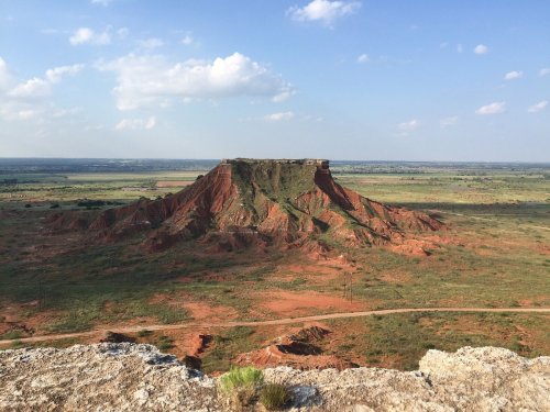 A scenic view of a red rock formation rising above a green landscape under a blue sky with scattered clouds.