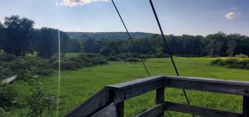 A scenic view from a wooden platform overlooking a lush green field and distant hills under a clear blue sky.