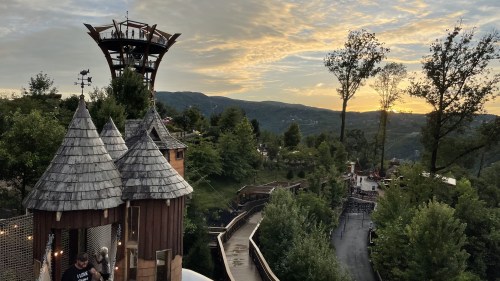 A scenic view of a wooden playground with towers, surrounded by trees and mountains at sunset.