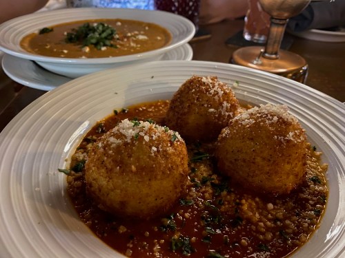 Three golden-brown balls on a bed of sauce, garnished with herbs and cheese, with a bowl of soup in the background.