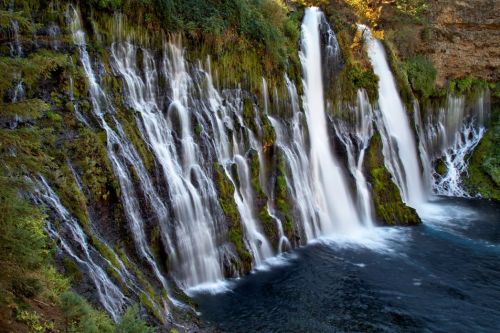 A serene waterfall cascades over moss-covered rocks into a deep blue pool, surrounded by lush greenery.