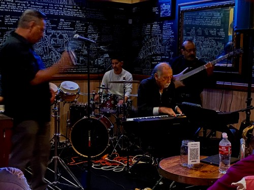 A jazz band performs on stage, featuring a pianist, drummer, and bassist in a lively venue with chalkboard decor.