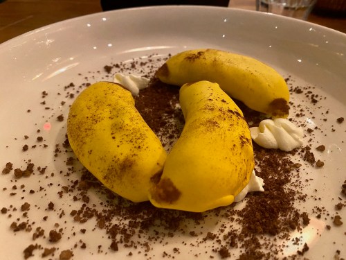 Three yellow dessert items shaped like bananas, sprinkled with cocoa powder and served with dollops of cream.