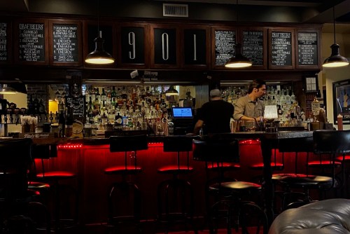 A dimly lit bar with a wooden counter, shelves of liquor, and two bartenders preparing drinks.