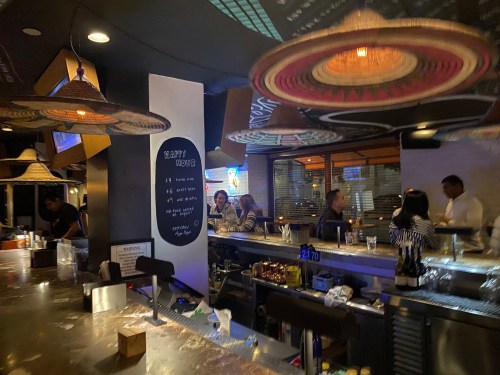 A lively bar scene with patrons chatting, colorful sombreros hanging, and a chalkboard menu in the background.