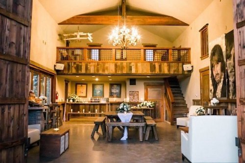 A rustic event space with wooden decor, a chandelier, and a cozy seating area, featuring a bar and staircase.