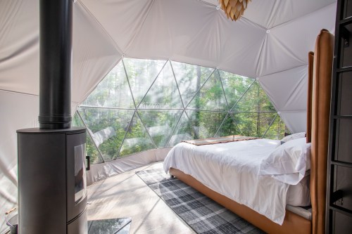 Cozy interior of a geodesic dome bedroom with a bed, wood stove, and large windows overlooking greenery.