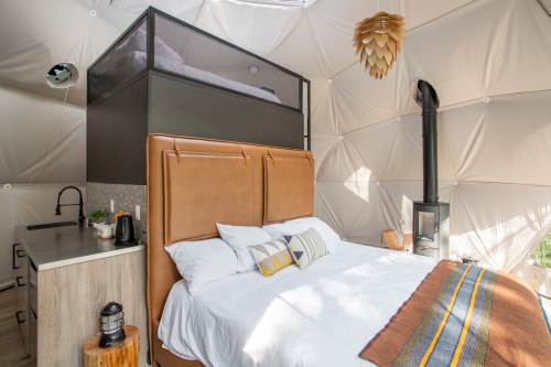 Cozy interior of a geodesic dome with a bed, kitchenette, and a lofted sleeping area above. Warm, modern decor.