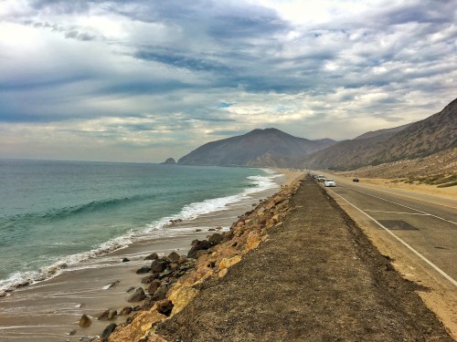 A scenic coastal road alongside a beach, with mountains in the background and gentle waves lapping at the shore.